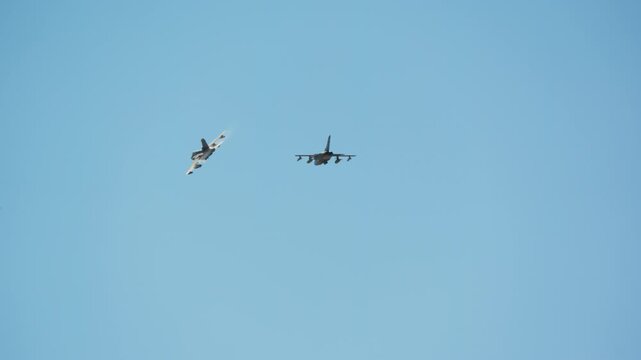 Two fighter jets execute a slow motion aerial maneuver against clear blue sky, one aircraft banking sharply while maintaining formation with the other, demonstrating coordinated flight