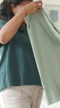 Vertical video: Picking up mature woman in dark teal top folding pale green T-shirt by bed, storing