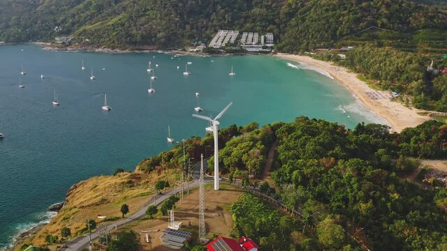 Aerial view panorama showing yachts anchored in the turquoise bay, a white wind turbine generating clean energy on a green headland, and the beautiful sandy Nai Harn beach, Thailand. Drone footage