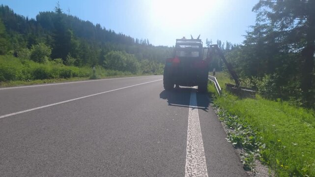 Pov driving behind a tractor with a flail mower cutting grass on the roadside on a sunny day