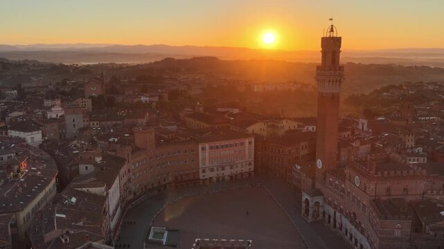 Tuscan city of Siena at sunrise, aerial view with the rising sun behind the famous medieval tower of Torre del Mangia, drone shot of piazza del Campo in Siena in the morning, travel in Tuscany, Italy