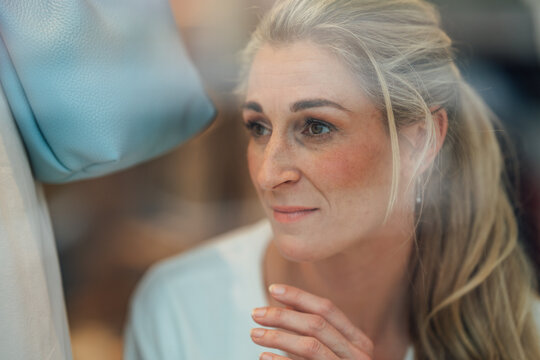 Portrait of an elegant blonde woman viewing a fashion store display through glass with reflections and sophisticated urban style.