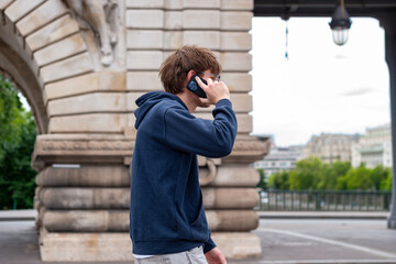 Fototapeta premium A young man stands by a bridge railing over a river while holding a smartphone. He adjusts his eyeglasses with his hand and looks down outdoors during the daytime.