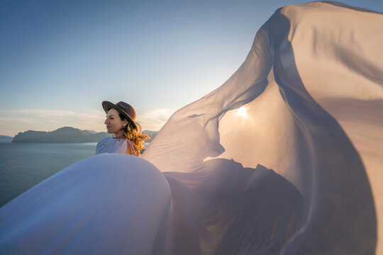 Woman hat dress, elegant female standing on cliff at sunset with fabric flowing in wind, inspirational nature lifestyle concept