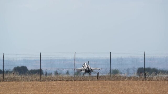 Single-engine fighter jet descending toward airfield, approaching landing zone with extended landing gear, passing over perimeter fencing and field boundary markers during daylight conditions
