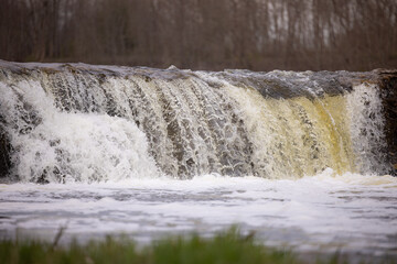 Kuldiga Venta River spring waterfall cascading over rocky ledge into calm water below, surrounded by sparse vegetation and trees in the background © Riga Frames