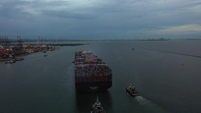 Large red container ship expertly maneuvered through darkened channel at bustling industrial port with precise tugboat guidance under overcast skies, highlighting maritime transport challenges.