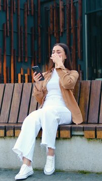 Frustrated young woman holding a smartphone, frowning and reacting to bad news, showing stress and disappointment while sitting on a bench outdoors near a modern office building. Slow motion
