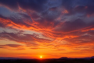 Fototapeta premium Sun dips below distant hills, painting sky in fiery hues. Crimson clouds swirl with deep purples and golds. Horizon glows warmly as light fades into dusk