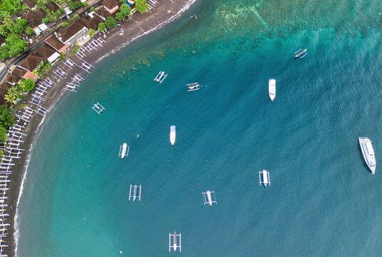 Aerial view of Jemeluk Beach with traditional outrigger boats floating on turquoise water near the dark volcanic sand shoreline in Kecamatan Abang, Bali, Indonesia.