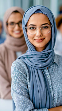 A woman wearing glasses and a blue hijab smiles at the camera