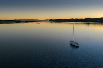 Obraz premium A lone sailboat rests on calm waters at sunset in Manukau Harbour, Auckland, New Zealand. The serene scene captures the peaceful end of a day on the Harbour.