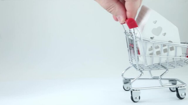 Woman hand slowly pushes small metal shopping cart with miniature white house model on white background, symbolizing real estate purchase and home sale concept.