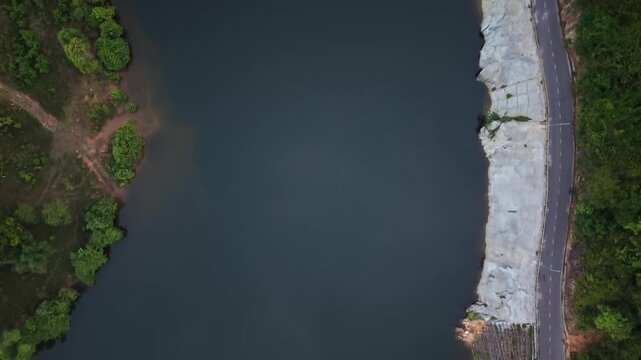 Cinematic top down drone flight over a tranquil river bordered by a winding mountain road and lush tropical vegetation in Vietnam. Aerial footage highlighting natural landscapes, infrastructure, and t