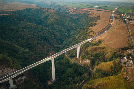 Aerial view of Puente Beltran spanning a deep, lush green canyon with a winding road below under soft golden hour light in Tonila, Jalisco, Mexico.