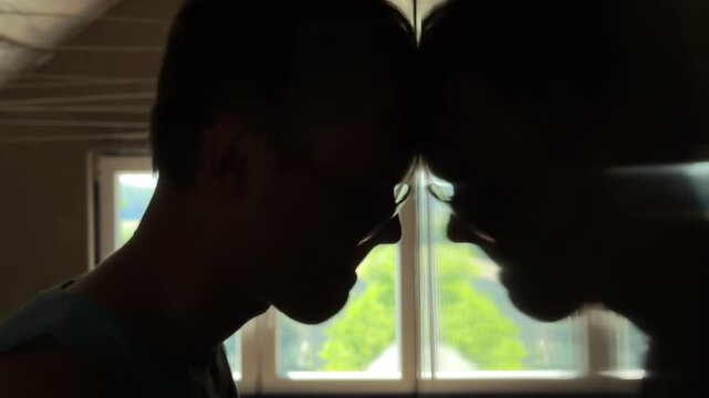 Close-up depressed teenager in a dark bedroom pressing his head against a mirror and staring at his reflection
