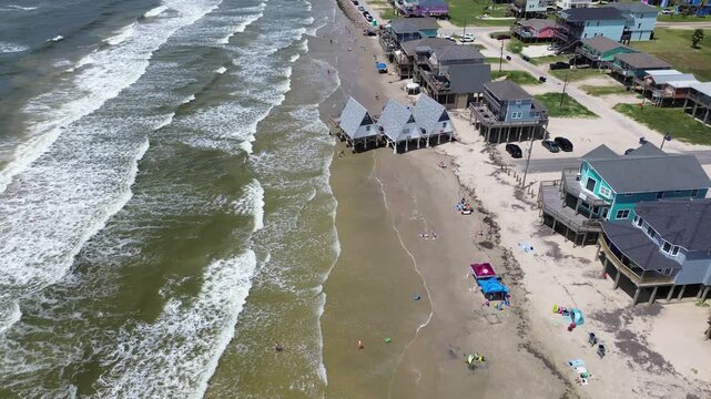 Iconic A frame beach structures stand near water, surrounded by multi story residential homes on elevated pilings. Families walk along expansive shoreline bright sky, Surfside Beach, Freeport, TX