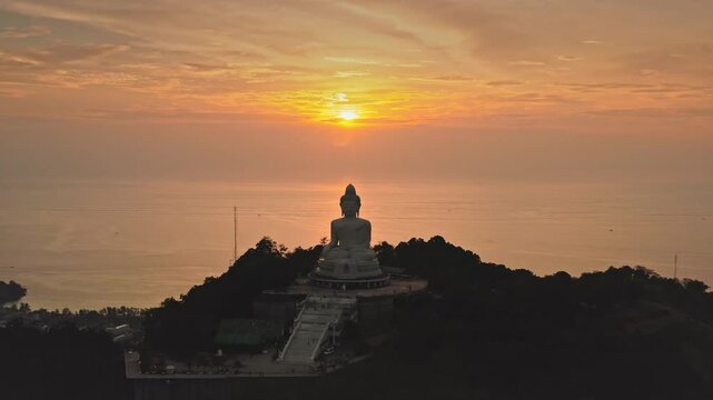 Big Buddha Phuket statue sitting peacefully on top of Nakkerd Hill, overlooking the coastline and ocean with a vibrant orange sunset creating a golden glow over the horizon. Aerial view drone flight