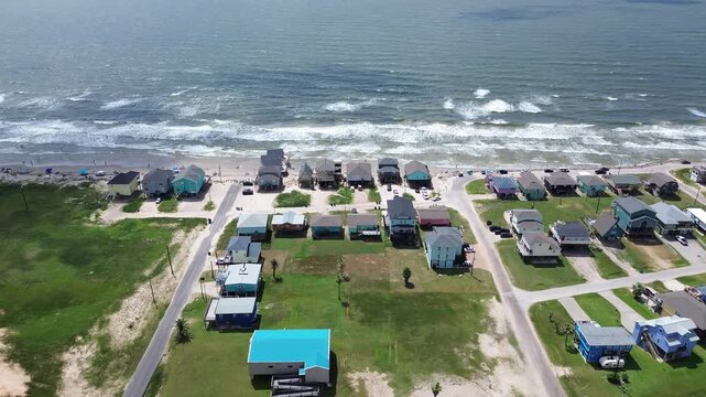 Multi level homes line beachfront near Starfish St, exhibiting pastel siding metal roofing. Green patches sit between stilted structures against white capped Gulf waters, Surfside Beach, Texas