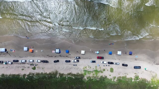 Vehicles park in organized rows along sandy beach near waterline. Numerous colorful tents dot sand while swimmers navigate rolling waves washing over wet shore, Surfside Beach, Freeport, Texas