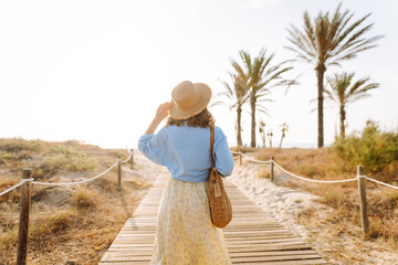 A beautiful woman in a straw hat walks along a wooden path toward the beach. A young woman enjoys...