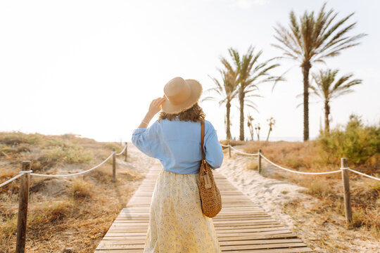 A beautiful woman in a straw hat walks along a wooden path toward the beach. A young woman enjoys the seascape at sunset. Concepts of beauty, nature, and lifestyle.