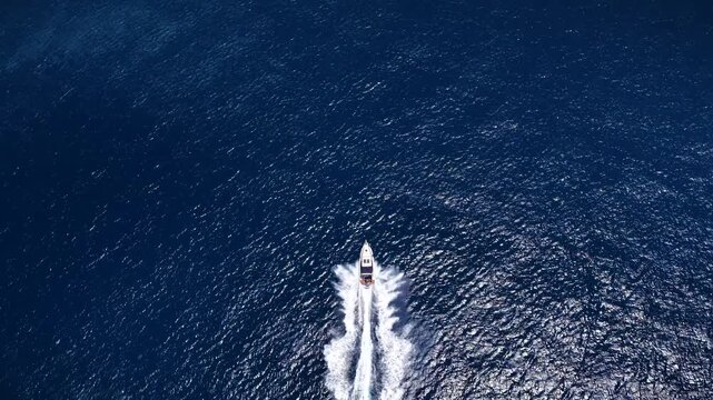 High aerial top view of a motor yacht cruising with high speed over blue, sparkling ocean and leaving a trail of waves and surf