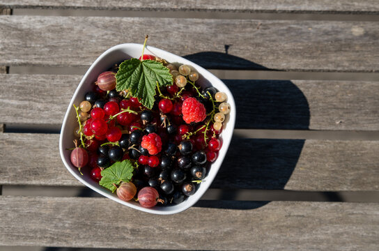 A mix of fresh tasty berries in a quadrangular white dish on a wooden table, two leaves in it, shadow on the right, dish on the left