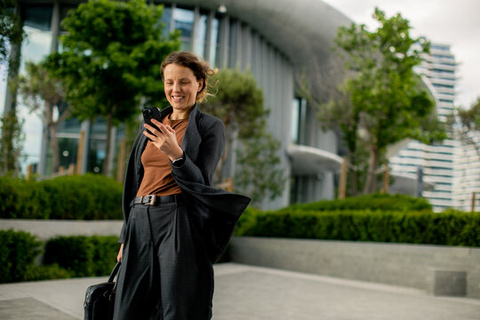 Woman smiles while looking at phone near modern building in the city