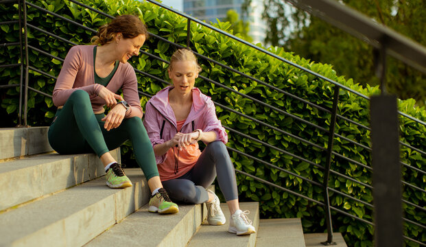 Two women sit on steps in a park and check fitness watches during a workout break
