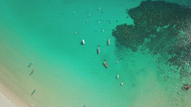 Tropical beach in Phuket Thailand featuring crystal clear turquoise water, white sand, and numerous traditional longtail boats floating among people snorkeling and exploring the vibrant coral reef