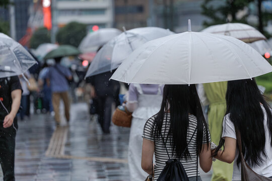 People walking with umbrellas on a rainy Nagoya street. Two friends share a white umbrella on wet pavement with a blurred urban background.