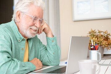 Fototapeta premium Mature adult man with white hair and beard smiling while working remotely on a laptop, enjoying online learning and modern technology from his home office