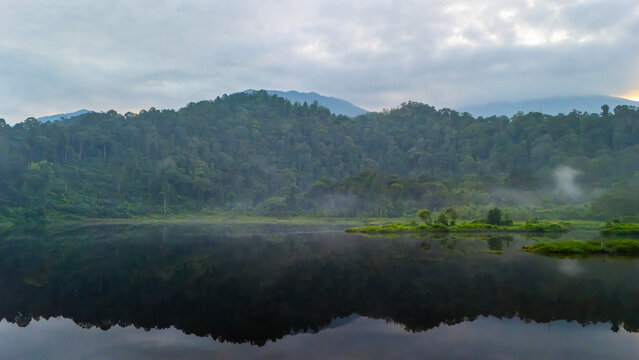 Aerial view of a serene lake surrounded by lush green mountains and dense forest under a cloudy sky with patches of morning mist in West Java, Indonesia.