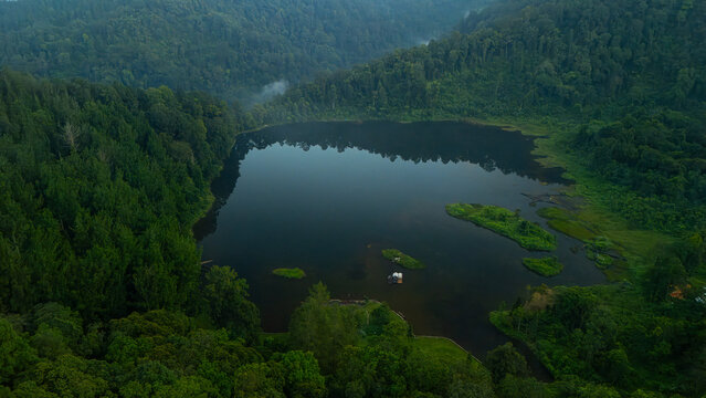 Aerial view of a serene lake nestled within a dense tropical forest, featuring calm dark waters, small grassy islets, and lush green hills under soft daylight in West Java, Indonesia.
