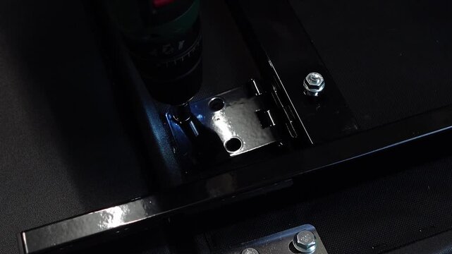 Hands using a cordless drill with a socket bit to tighten bolts into a black metal hinge during furniture assembly