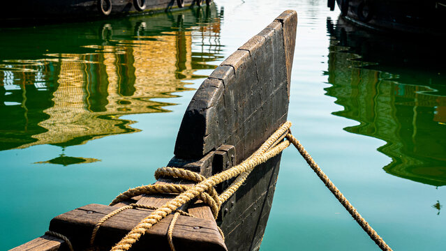 Closeup view of a traditional wooden gondola prow with a rope tied, reflected in the green water of a canal