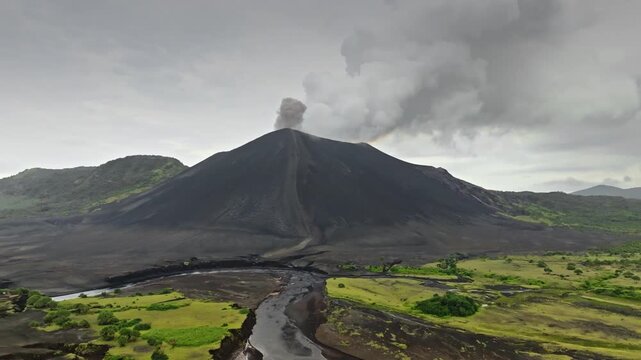 Mount Yasur volcano on Tanna Island, Vanuatu, actively erupting and sending ash and smoke into cloudy sky, with a dark landscape and lush green vegetation in the foreground. Aerial view drone panorama