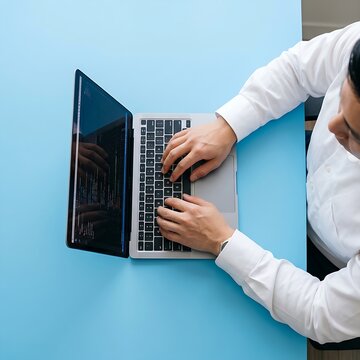 A man wearing a white dress shirt operates a laptop computer placed on a vivid blue background.