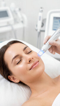 Smiling young Caucasian woman receiving a professional facial hydro-peeling skin treatment at a beauty clinic. Cosmetologist hand in glove holding a modern blue light laser device.