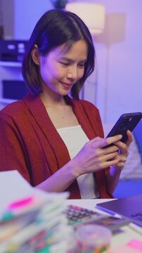 Smiling young Asian businesswoman using smartphone while wearing headset in office.