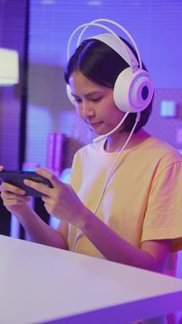 Excited young Asian girl playing mobile game on smartphone and headset at home.