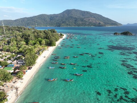 Aerial view of Koh Lipe Pier with long-tail boats anchored in turquoise waters along a sandy beach and lush vegetation in Tambon Ko Tarutao, Chang Wat Satun, Thailand.