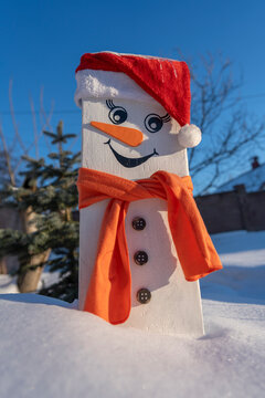 Handmade wooden snowman standing in winter snow. Creative DIY wooden board snowman wearing red Santa hat and orange scarf placed in deep snow under blue sky for rustic Christmas outdoor decoration