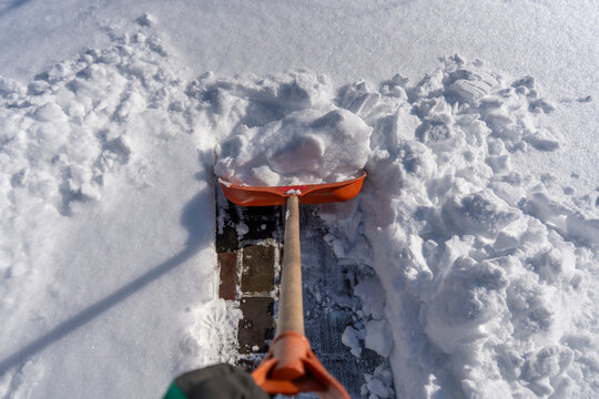 Clearing snow from brick pathway with orange shovel. POV view of shoveling fresh winter snow from brick pavement with an orange snow shovel during sunny day representing seasonal house maintenance