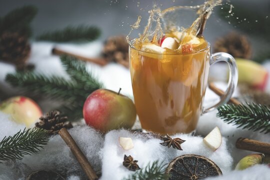 Apple punch splashes out of a glass cup with apple pieces, surrounded by cinnamon sticks and pine branches in a wintry environment