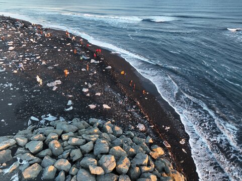 Aerial view of Diamond Beach with ice chunks on black volcanic sand, waves crashing on the shore, and people walking along the coastline in Hofn, Sveitarfelagid Hornaf, Iceland.