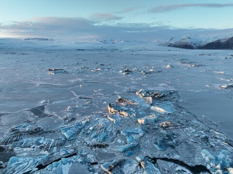 Aerial view of Diamond Beach featuring scattered ice chunks on the frozen shoreline with snow-covered mountains in the background under a soft winter sky in Hofn, Sveitarfelagid Hornaf, Iceland.