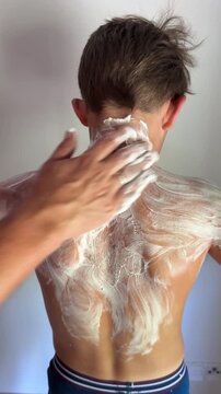 Sunburned boy at home applying ointment to back, back view