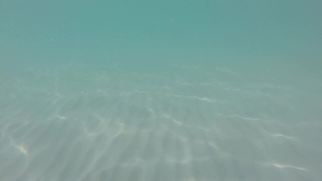 Underwater view of a rippled sandy seabed beneath crystal clear turquoise water with shimmering sunlight caustics